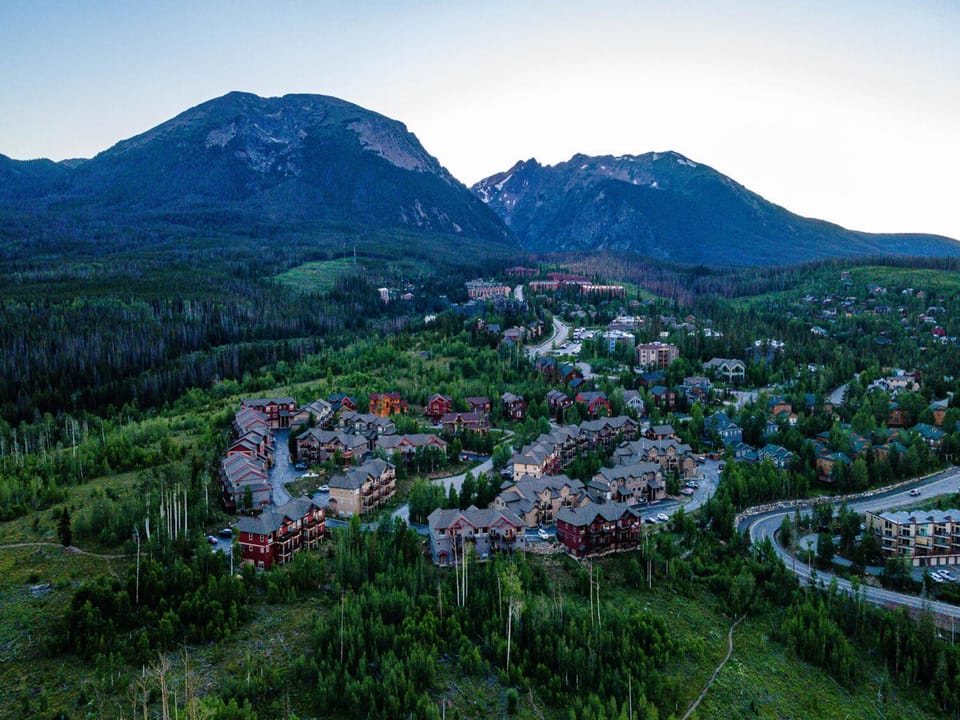 Aerial view of a mountain resort community with numerous buildings nestled among trees at the base of large, forested mountains under an evening sky.
