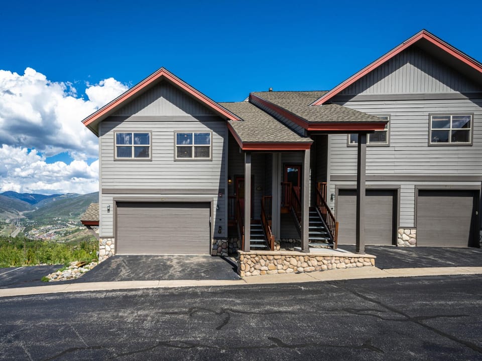 A two-story house with grey siding and a two-car garage, situated on a paved road. The front has a porch with steps, and scenic mountainous landscape is visible in the background under a clear blue sky.