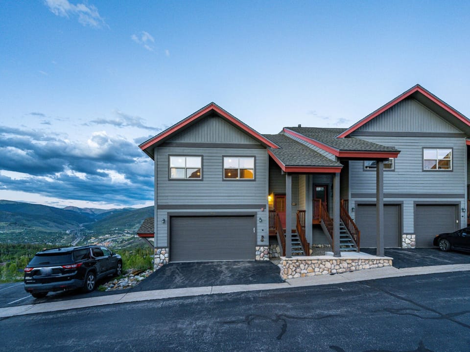 Duplex with two garages and a shared front staircase, located on a sloped street, facing a scenic valley and mountains under a partly cloudy sky.