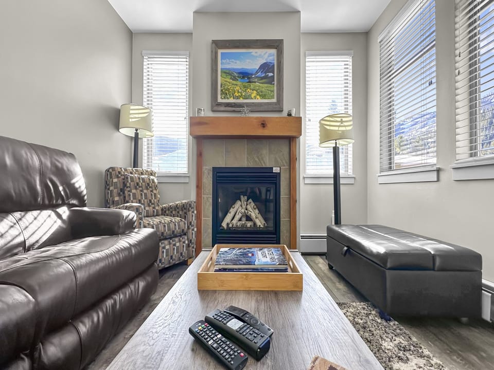 A living room featuring a fireplace with art above it, leather couch, patterned armchair, ottoman, and coffee table with a tray holding remotes and magazines. Sunlight streams in through windows on both sides.