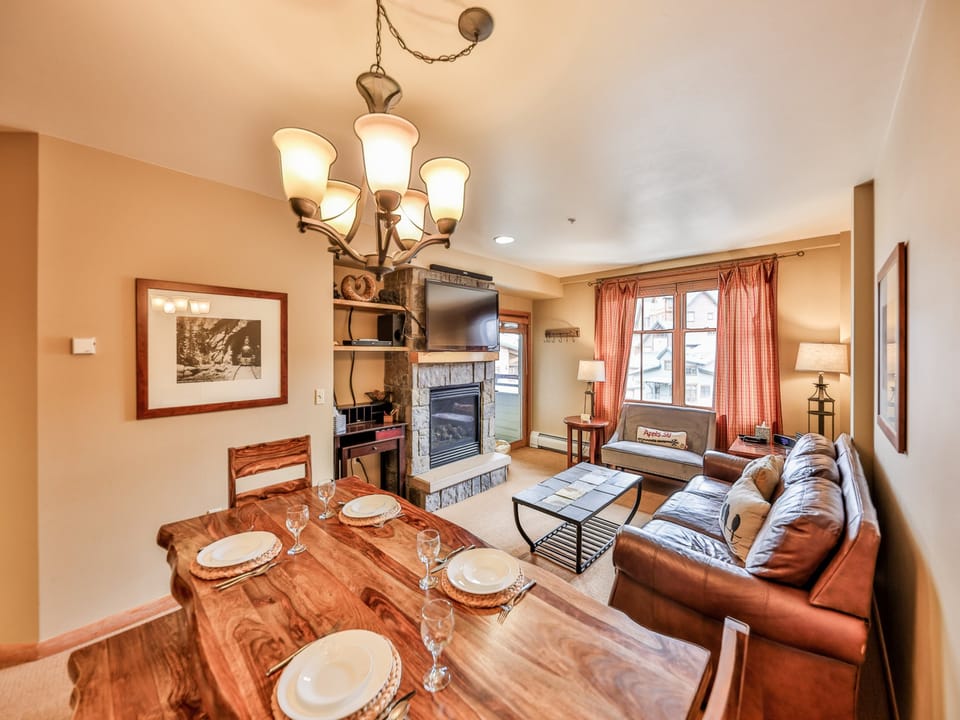 A well-lit living room with a dining table set for four, a leather couch, a TV above a fireplace, and large windows with orange curtains. Warm tones dominate the decor.