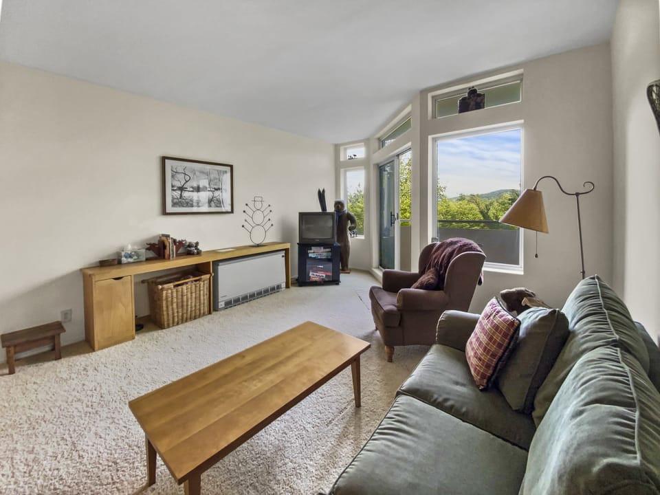 A living room with a couch, coffee table, armchair, and TV. There is a large window with outside view, framed artwork on the wall, and various items on a long wooden cabinet.