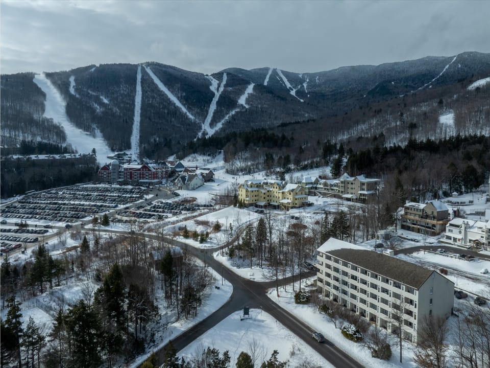 Aerial view of a ski resort village nestled in snow-covered mountains with multiple ski slopes, lodges, and parking areas during winter.