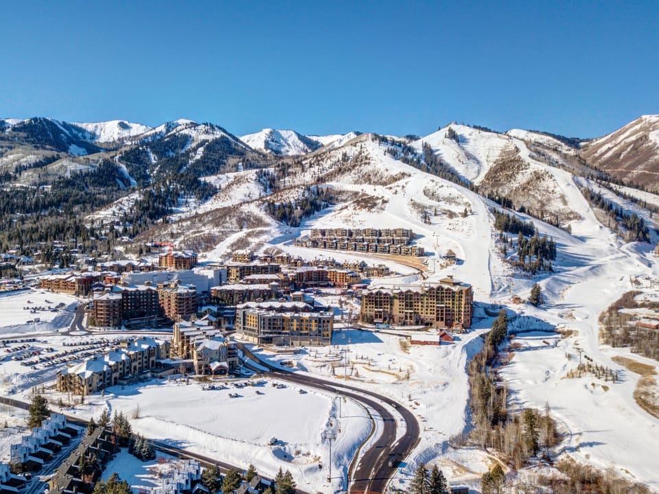 Aerial view of a snowy mountain resort with multiple buildings, ski slopes, and winding roads under a clear blue sky.