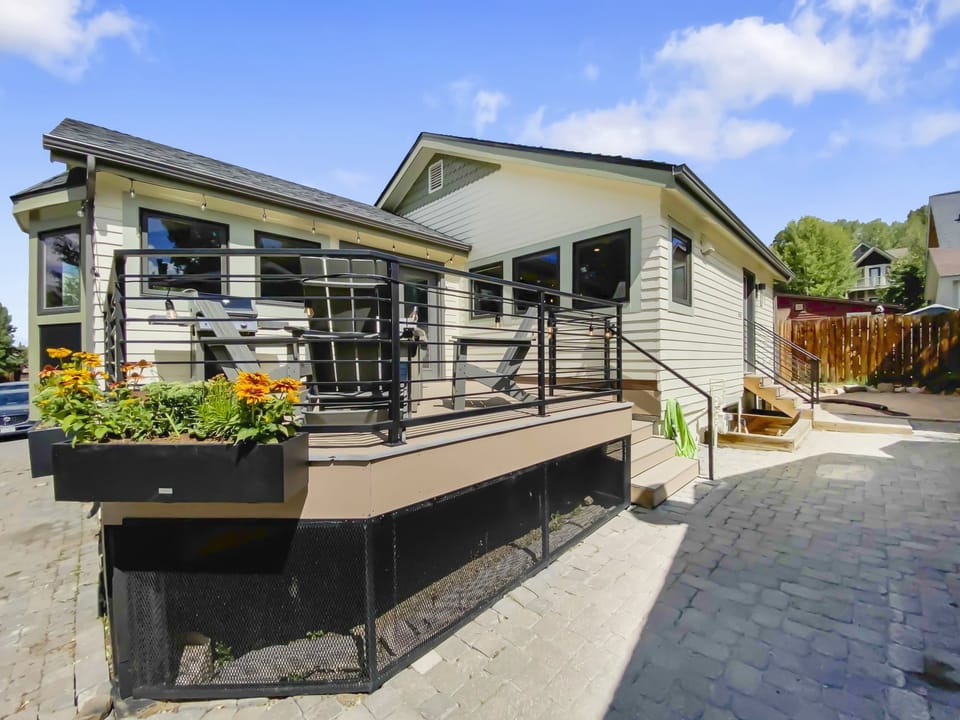 A modern house with a light exterior features a raised deck, metal railings, and flower boxes. The yard has a stone-paved walkway and steps leading to an entrance.