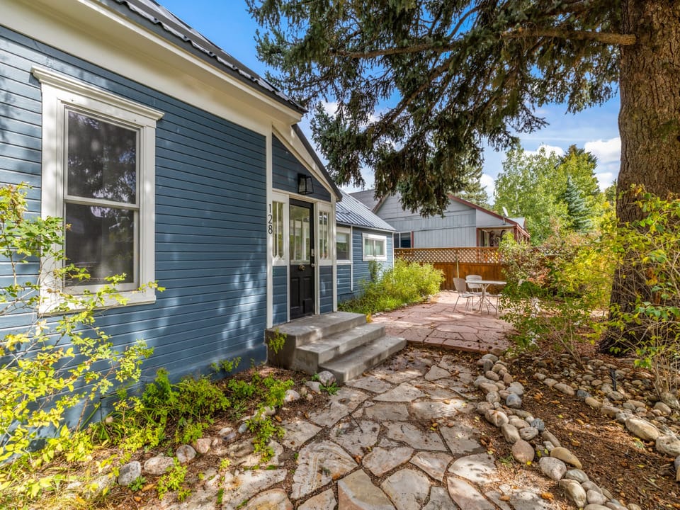 A charming blue cottage with inviting stone pathway through lush greenery and mature trees, leading to a quiet neighborhood setting perfect for your peaceful getaway.