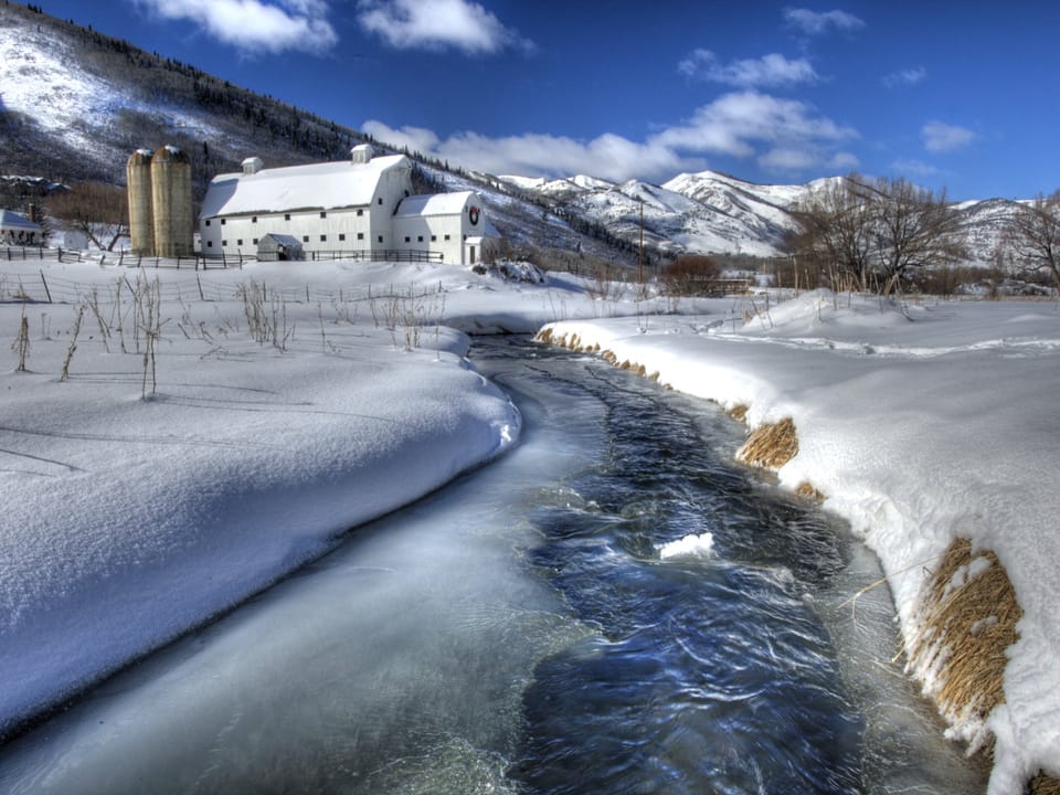 A snowy landscape with a barn, grain silos, and mountains in the background. A partially frozen stream runs through the scene under a mostly clear sky.