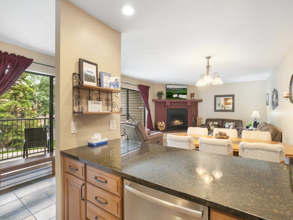 A kitchen with a granite countertop island overlooks a cozy living room featuring a fireplace, a mounted TV, a brown sectional sofa, a dining table with chairs, and a sliding glass door to a balcony.