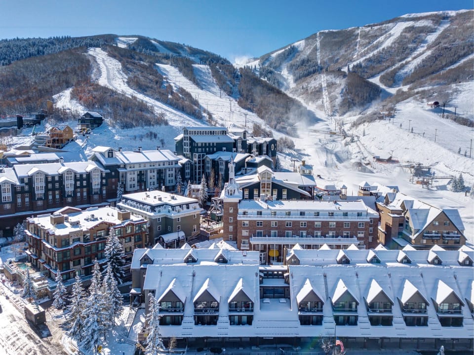 Aerial view of a snowy mountain ski resort with multiple buildings and ski slopes visible under a clear blue sky.