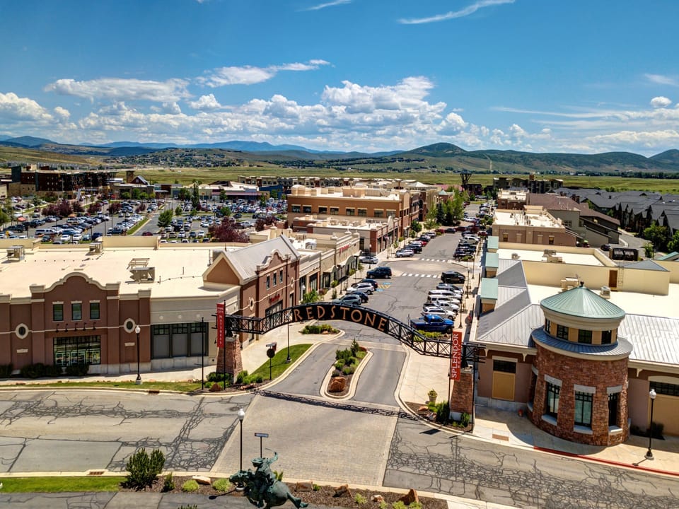 Aerial view of a commercial shopping district named Redstone, featuring retail buildings, parking lots, and a surrounding landscape of green hills under a partly cloudy sky.