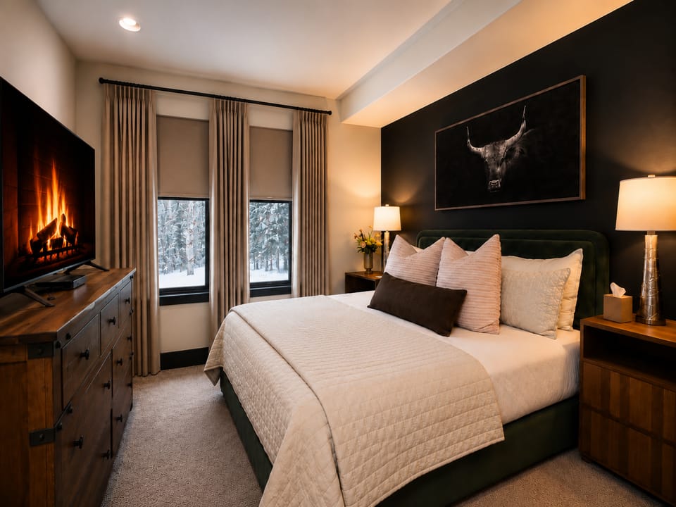 Cozy elegance awaits in your bedroom, featuring a plush bed dressed in soft linens, a glowing fireplace to warm chilly evenings, and windows framing a peaceful winter forest view.