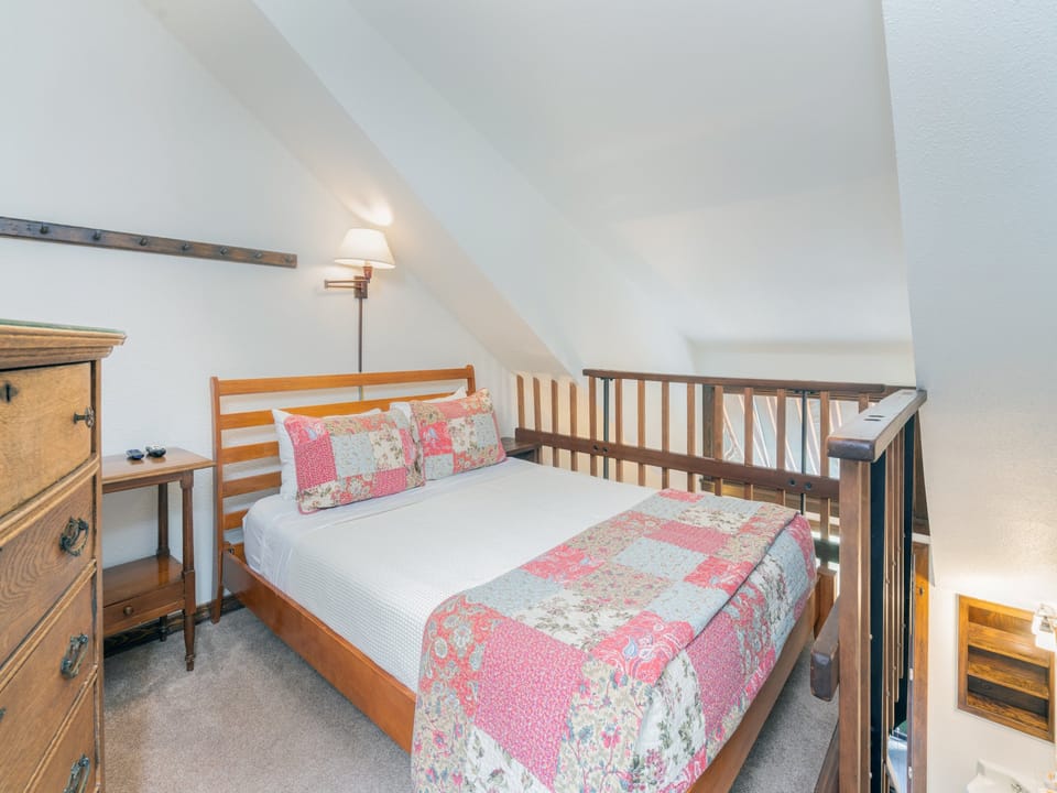 Your charming loft bedroom awaits with a cozy bed dressed in floral linens, warm lighting, and rustic wood furnishings. Perfect for a restful night's sleep.