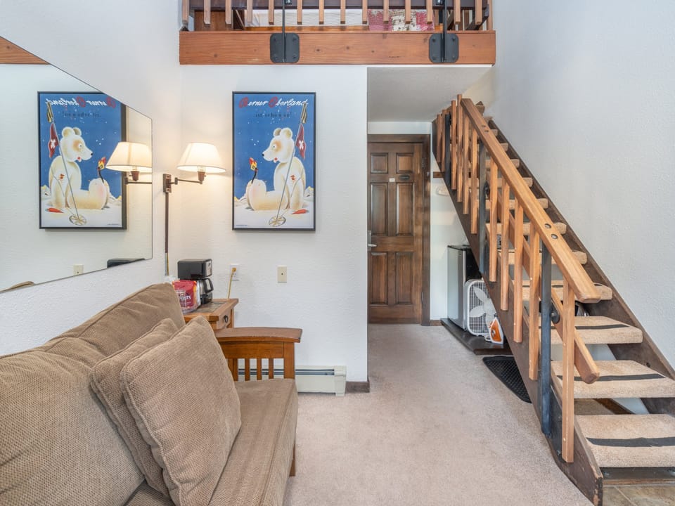 Your charming loft retreat features cozy living space with comfortable seating, warm lighting, and whimsical alpine artwork. A handcrafted wooden staircase leads to your private sleeping loft above, creating a unique two-story layout perfect for relaxing m