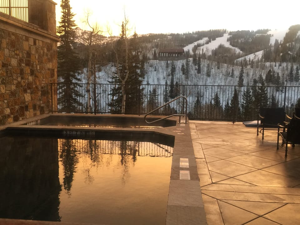 An outdoor pool and hot tub overlook snow-covered ski slopes framed by evergreen forests, with winter's golden hour lighting the mountain landscape.