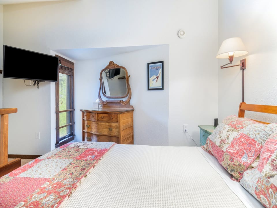 Your cozy bedroom awaits with a plush bed, charming vintage dresser, flat-screen TV, and bedside lighting. Natural light streams through the window, creating a warm and inviting retreat for your stay.