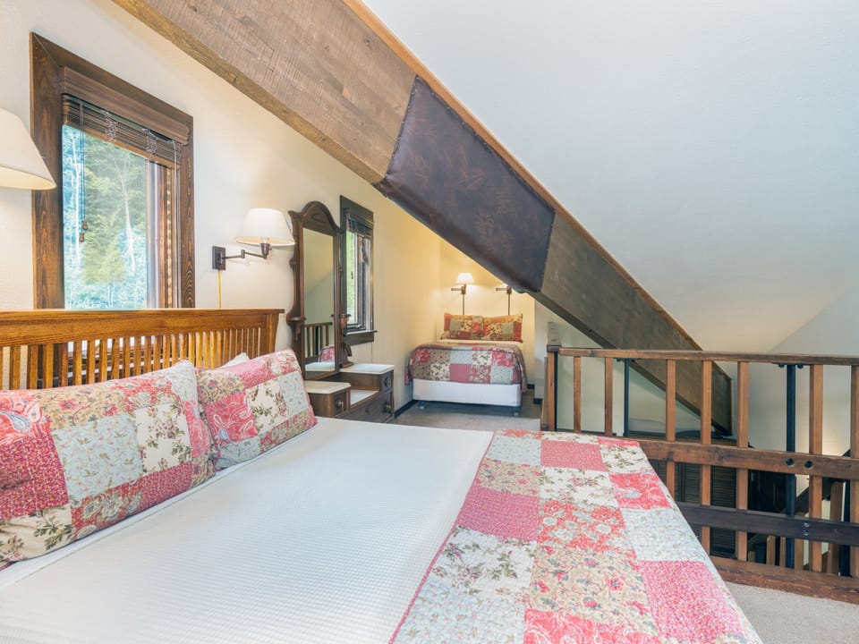 Your cozy loft bedroom features charming exposed beams, crisp linens with soft quilted accents, and warm natural light streaming through rustic wood-framed windows—perfect for restful mountain mornings.