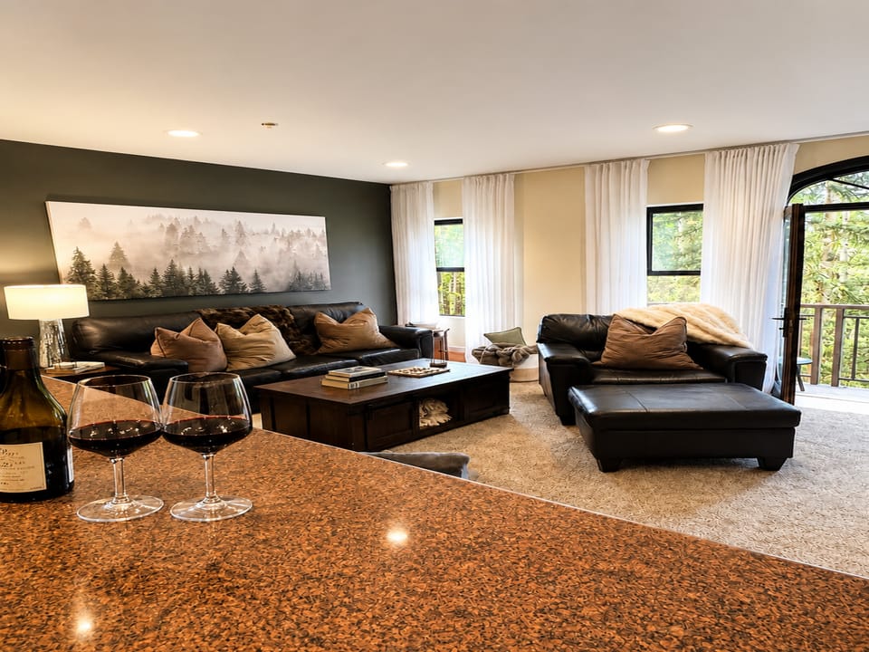 Unwind in your stylish living area with rich leather seating, a cozy throw, and a glass of wine waiting. Through the balcony door, lush greenery frames your peaceful retreat.