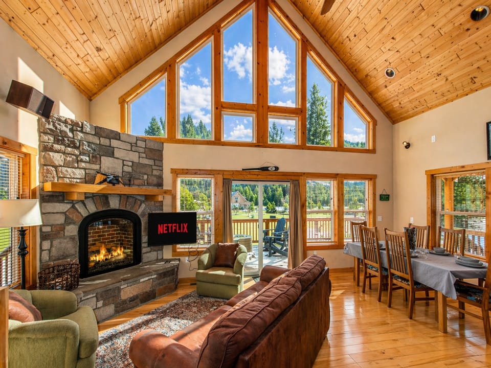Vaulted cabin living with a glowing fireplace and peaceful forest views