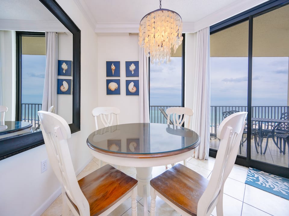 Dining area features a round table with four chairs, framed seashell art, and beachfront balcony
