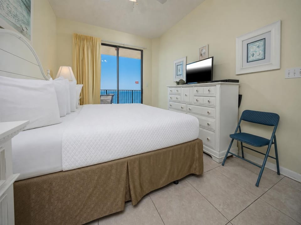 Bright master bedroom featuring a king bed, dresser, TV, and sliding glass doors leading to a balcony with a beach view