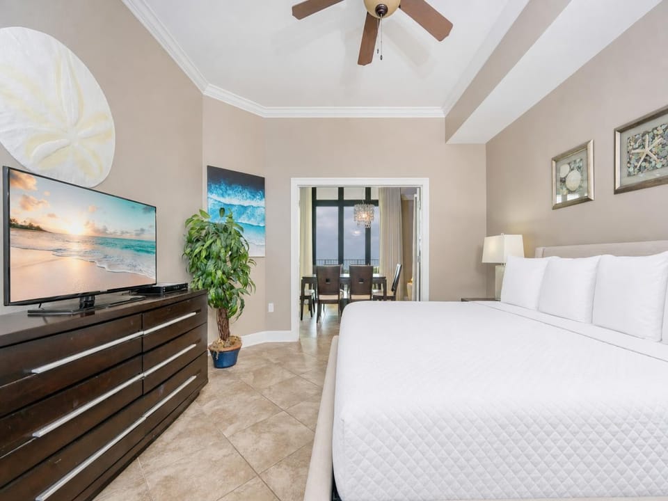 Relaxing master bedroom with a cozy king bed, sleek TV, and a view of the dining area and beautiful coastal artwork
