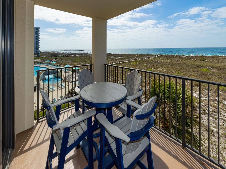 Balcony features a round table and four high-backed chairs, offering a view of the beach and gentle waves beyond