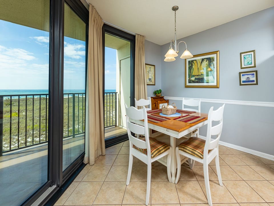 Bright dining area features a wooden table with colorful placemats, surrounded by white chairs, with a view of the beach