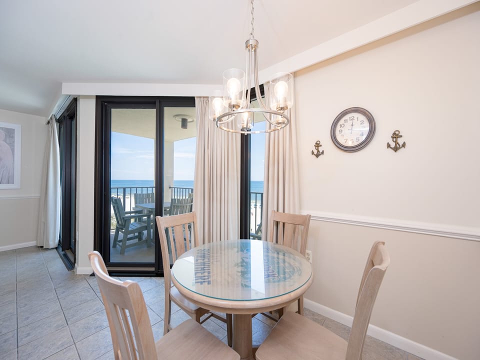 A round glass-top dining table with wooden chairs offers a view of the beach through large sliding glass doors