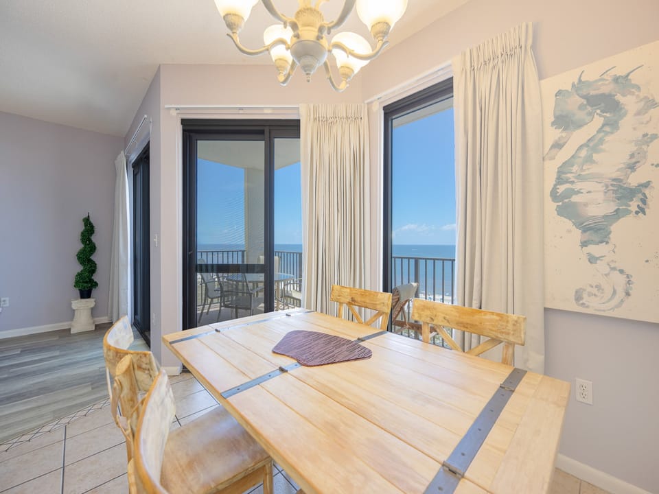 Dining area features a wooden table with four chairs, large windows showcasing a beachfront view, and coastal decor