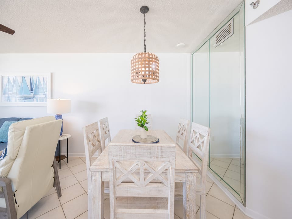 Bright dining area featuring a table surrounded by white chairs, with a beach view visible through large windows