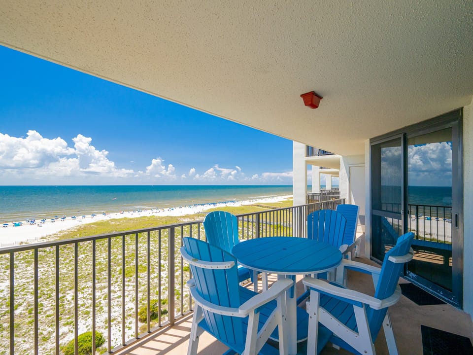 Bright blue chairs surround a circular table on the balcony, offering a stunning view of the glistening gulf and beach below