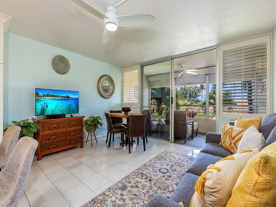 Unwind in your cozy living space where tropical breezes drift through open glass doors, framing lush palms and sunshine. Curl up on the plush sofa with cheerful accents, and let island life settle in around you.