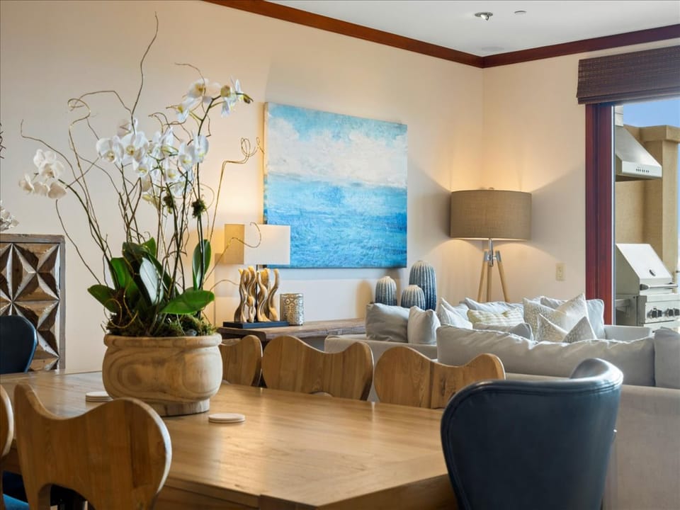 Gather around the elegant dining table, where coastal-inspired artwork and plush seating create your perfect space for memorable meals. Natural light spills through the window, while soft orchids add a welcoming touch.