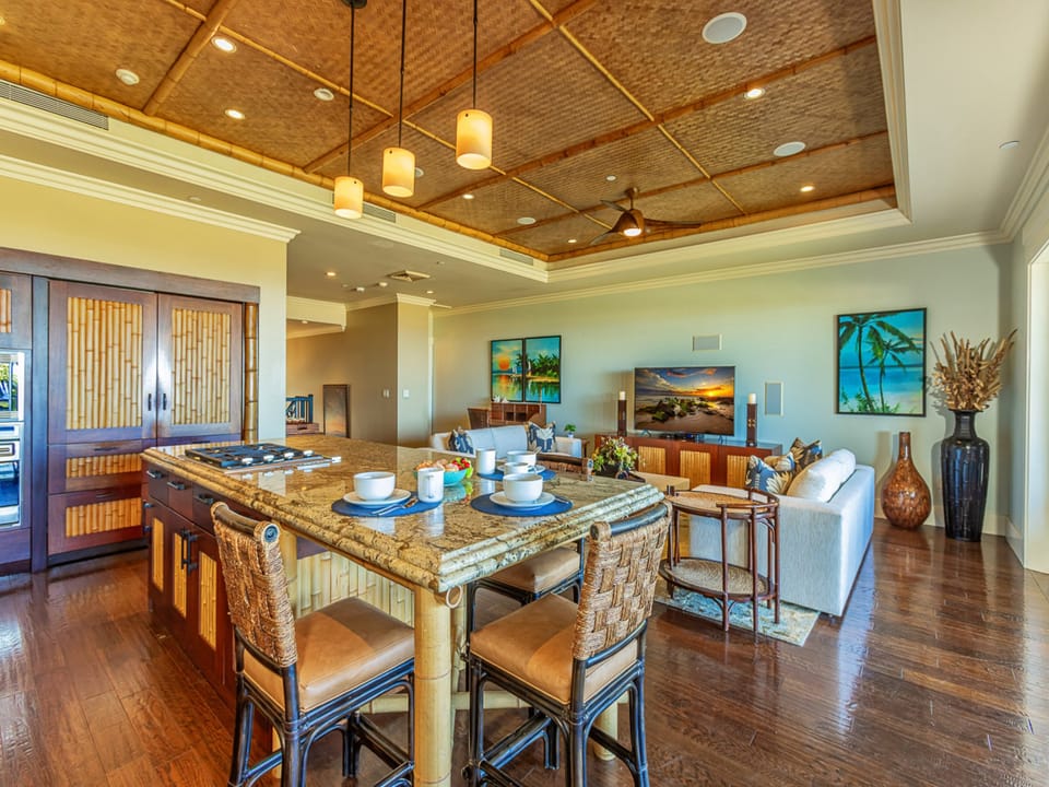 Step into your open-concept living space where island seating flows seamlessly into the lounge. Rich wood floors and a stunning woven bamboo ceiling create warmth, while tropical accents and beach-inspired art set a relaxed island tone throughout.