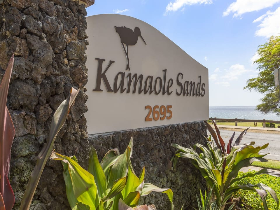 Kamaole Sands welcomes guests with a distinctive volcanic rock entrance marker set against blue sky and sparkling ocean views, flanked by tropical greenery.
