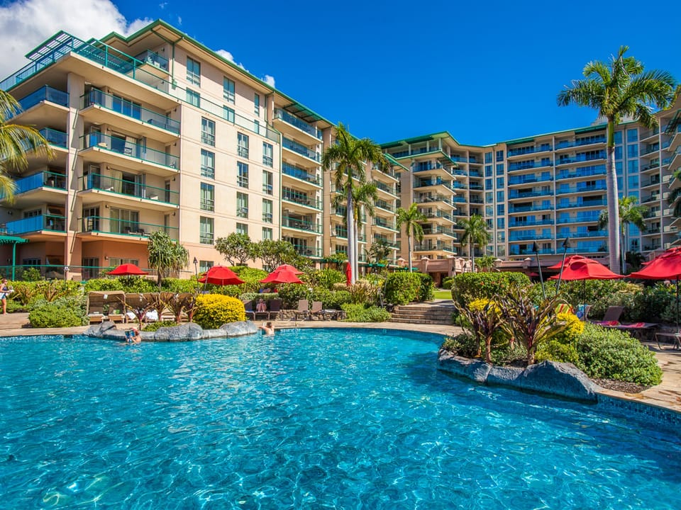 This resort-style pool winds through lush tropical landscaping, with sun loungers and red umbrellas beneath swaying palms. Multiple building wings surround the area, offering a bright, relaxing vacation setting.