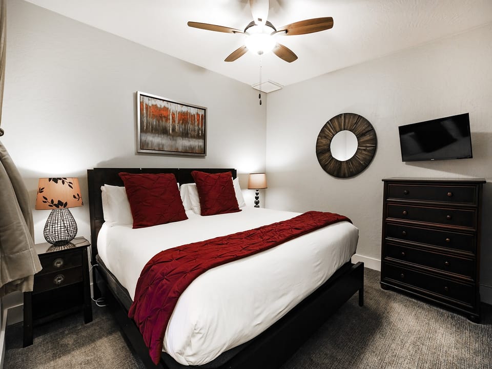 Guest bedroom with king bed, white linens and duvet and red accent runner and throw pillows, wall mounted TV and wooden dresser