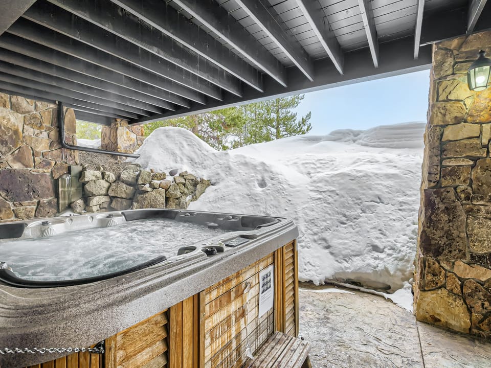 A hot tub under a wooden deck roof, surrounded by snow-covered stone walls and a snowbank in the background. A light fixture is attached to the stone wall on the right.
