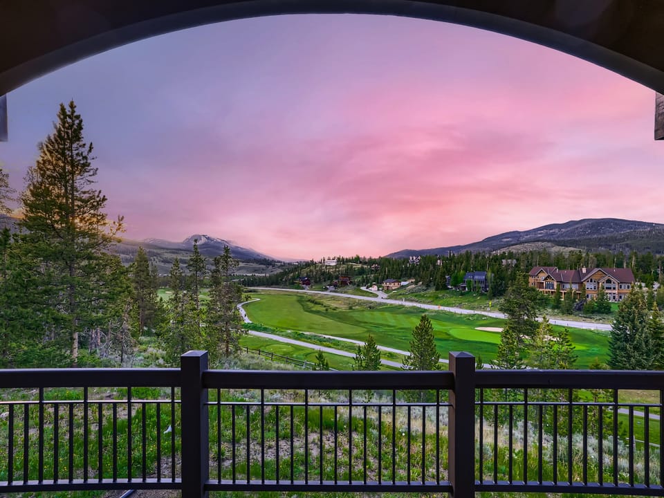 View of a scenic landscape featuring a golf course, trees, and distant mountains under a pink and purple sunset, seen through a balcony railing.