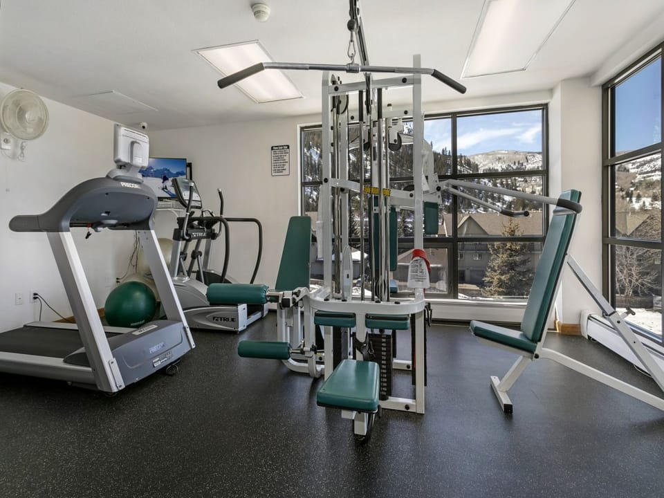 A well-lit gym with a treadmill, elliptical machine, seated weight machine, and stability ball placed near windows overlooking snow-covered trees and buildings.