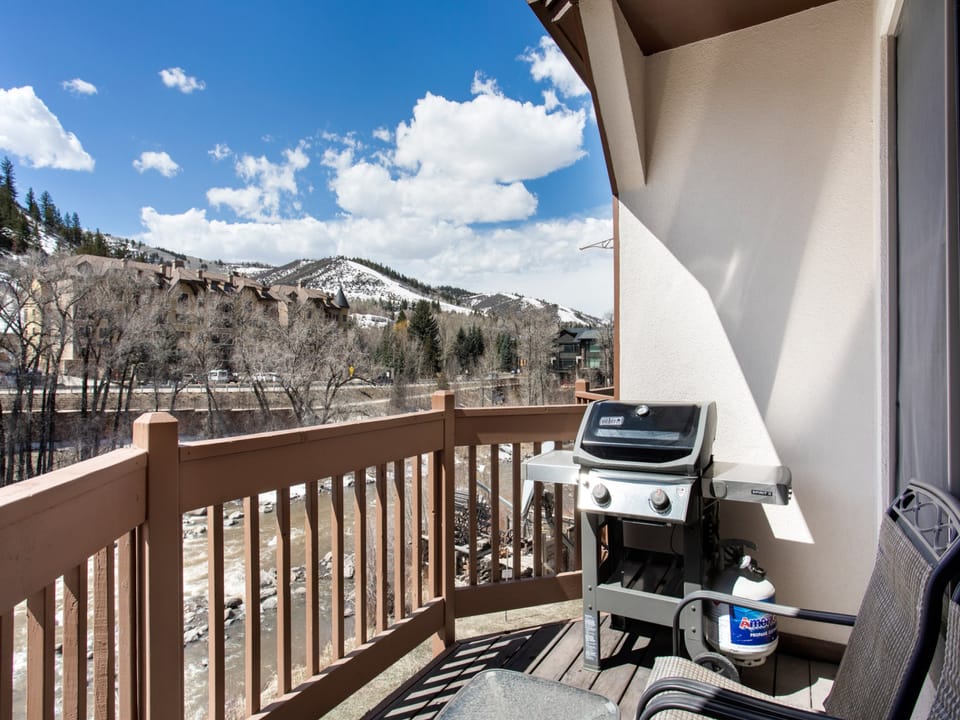 A wooden balcony with a gas grill and a chair overlooks a snow-covered mountainous landscape under a partly cloudy sky.