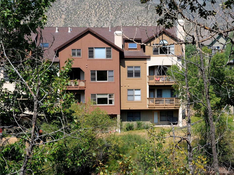 A multi-story residential building with brown and beige exterior, balconies, and windows, surrounded by trees and vegetation. Mountains are visible in the background.