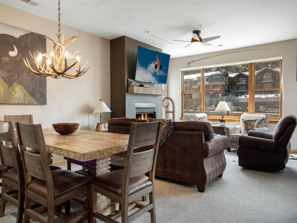 Modern living and dining room with a rustic wooden table, leather sofas, an antler chandelier, a fireplace, and a large window letting in natural light. A TV is mounted above the fireplace.