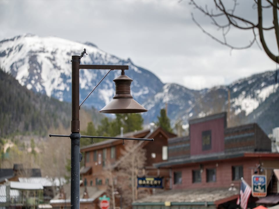 A street lamp in a small town with wooden buildings, snow-capped mountains, and trees in the background under a cloudy sky.