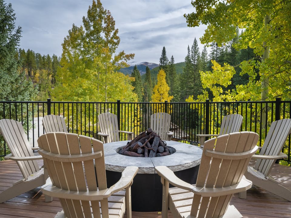 Eight wooden chairs encircle a fire pit on a deck surrounded by a forest with autumn trees, against a backdrop of mountains.