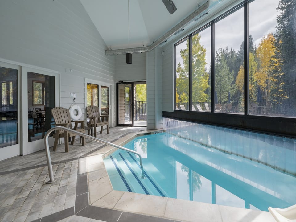 Indoor swimming pool area with large windows and a forest view. Features a metal handrail, wooden chairs, and a poolside seating area. Bright natural light floods the room through the windows.