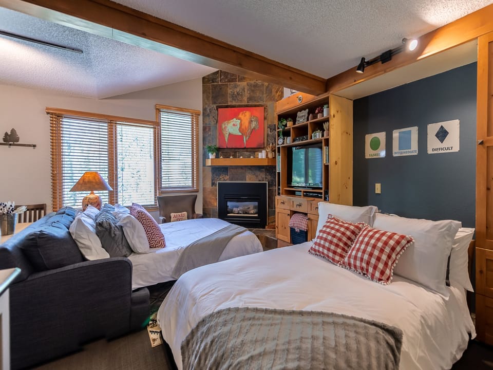 A cozy bedroom with two beds, a stone fireplace, a TV, a couch, and large windows. The beds have white linens and decorative pillows. The walls have various artworks and wooden accents.