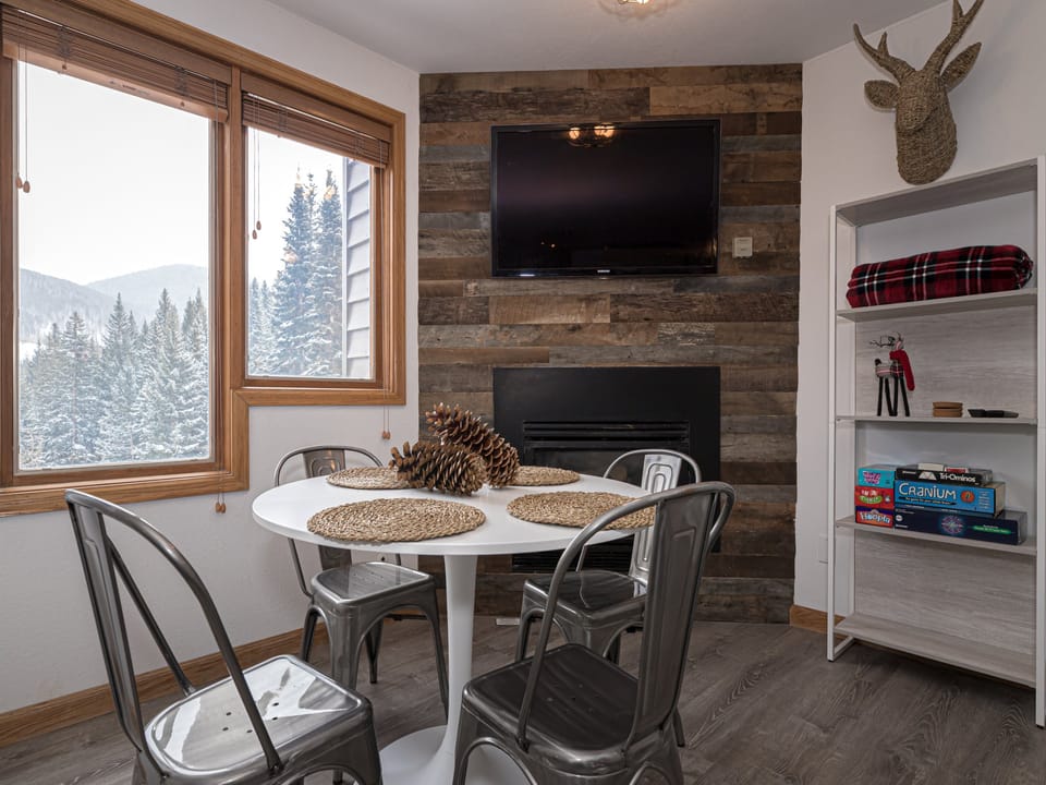 A small dining area with a round table and four metal chairs. The table has a pinecone centerpiece and placemats. A mounted TV and fireplace are on the wood-paneled wall. Snowy landscape visible through the window.