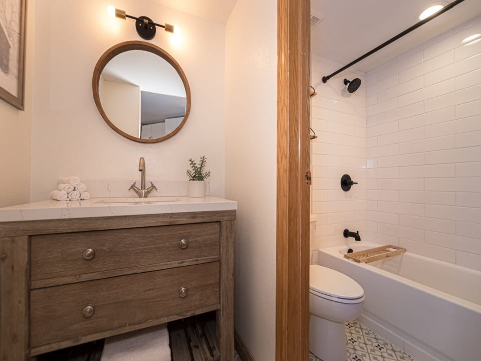 A bathroom featuring a wooden vanity with a round mirror and sink, a potted plant, a white-tiled bathtub with black fixtures, and a folded towel on the sink.