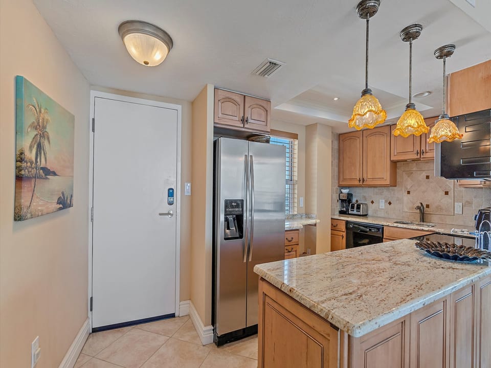 A modern kitchen with wooden cabinets, granite countertops, stainless steel appliances, pendant lights, and a wall painting of a tropical scene. An exterior door is on the left side of the image.